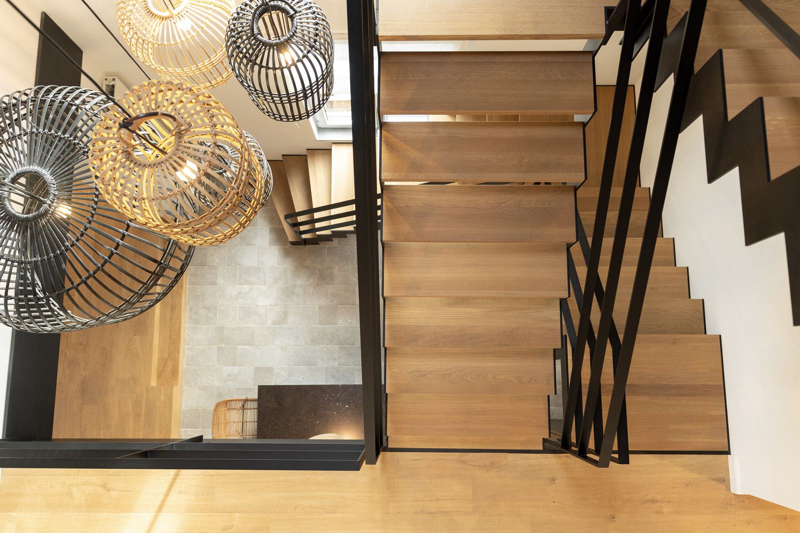 Houten trap met zwarte stalen balustrade van bovenaf, met rieten hanglampen