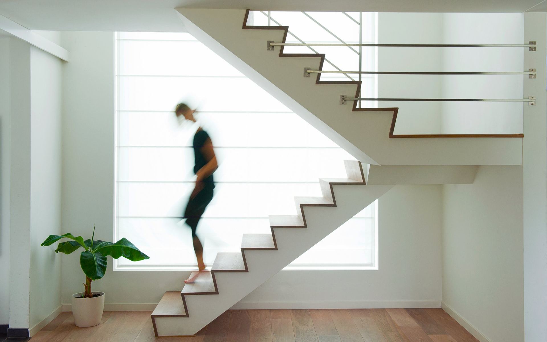 Iemand loopt een witte Z-trap met houten treden af, voor een groot raam